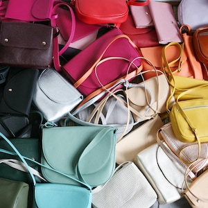 May include: A collection of colourful leather crossbody bags in various shades of blue, green, yellow, pink, red, and silver. The bags are arranged in a pile, showcasing their different shapes and sizes.