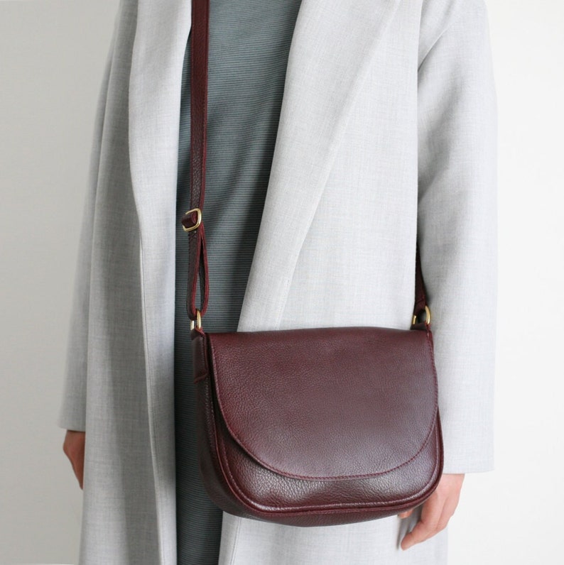 Pu&ograve; includere: Una borsa a tracolla in pelle color bordeaux con una fibbia dorata e una lunga tracolla. La borsa &egrave; indossata sopra un blazer grigio.