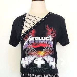 May include: Black t-shirt with a graphic of a hand holding strings attached to crosses. The text "Metallica" and "Master of Puppets" are printed on the shirt. The shirt has a lace-up detail on the neckline.