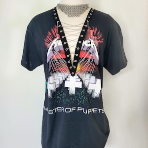 May include: Black t-shirt with a graphic of the band Metallica's "Master of Puppets" album cover. The shirt has a lace-up detail at the neckline with silver chain accents.