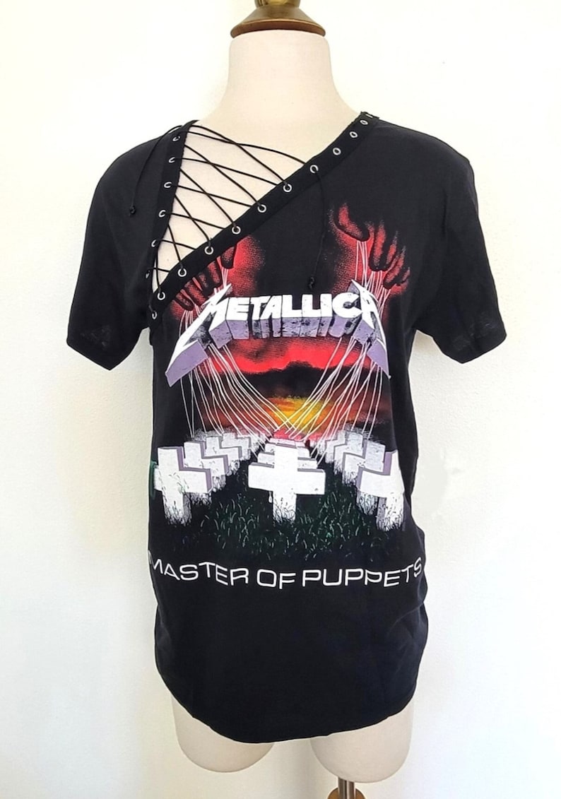 May include: Black t-shirt with a graphic of a hand holding strings attached to crosses. The text "Metallica" and "Master of Puppets" are printed on the shirt. The shirt has a lace-up detail on the neckline.