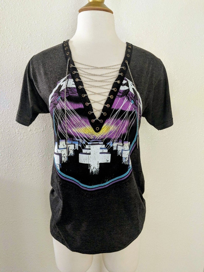 May include: A black t-shirt with a graphic print of a purple and blue design with white crosses. The shirt has a lace-up detail at the neckline with silver chains.