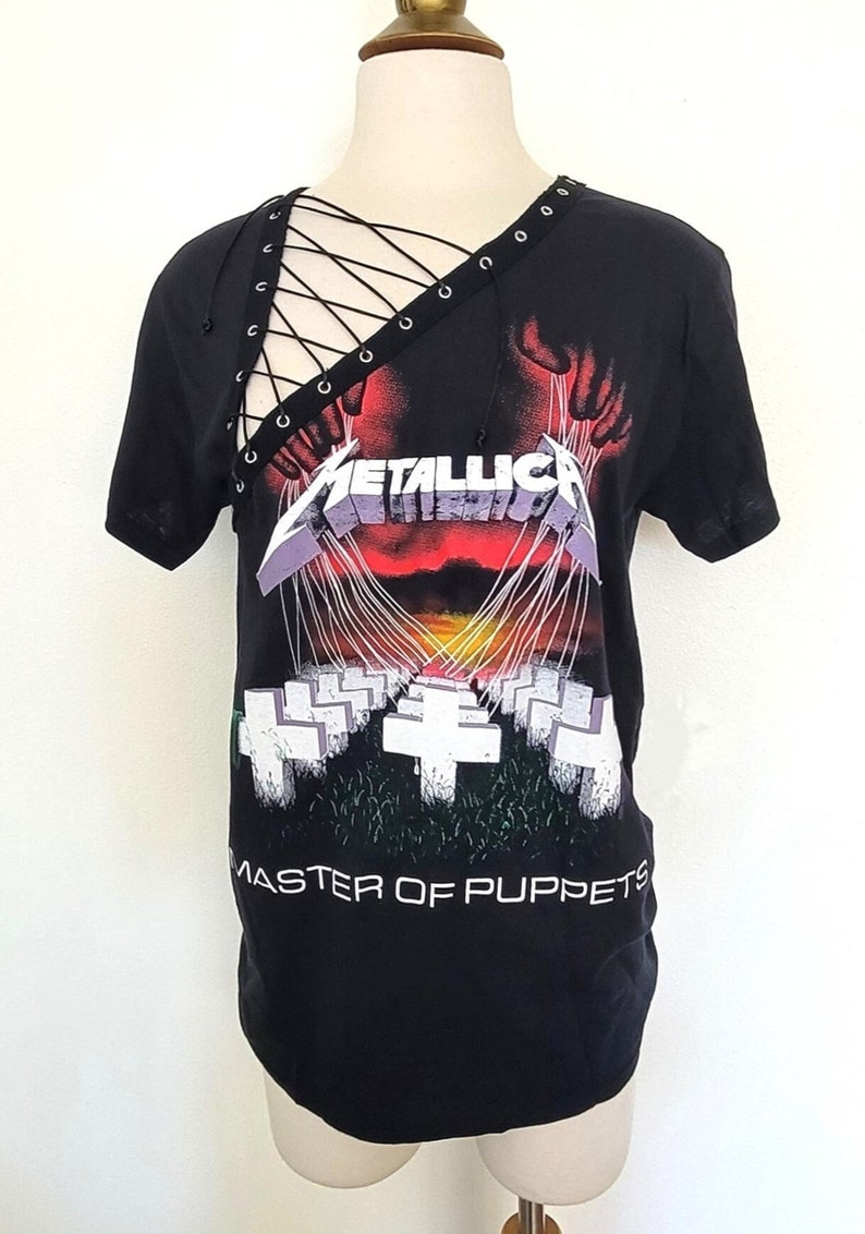 May include: Black t-shirt with a graphic of the band Metallica's "Master of Puppets" album cover. The shirt has a lace-up detail at the neckline.