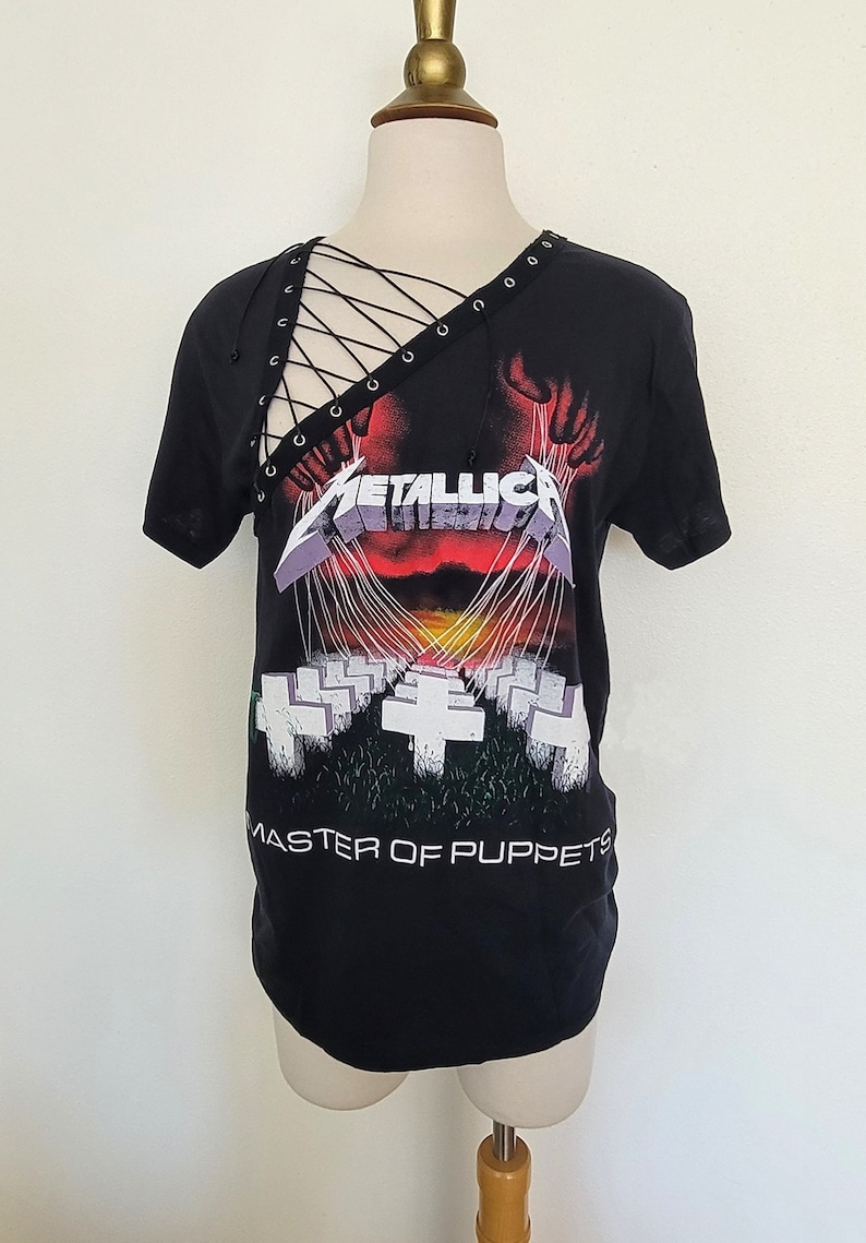 May include: Black t-shirt with a graphic print of the band Metallica's "Master of Puppets" album cover. The shirt has a lace-up detail at the neckline.