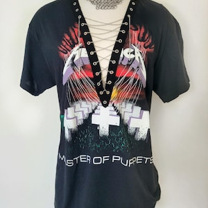 May include: Black t-shirt with a graphic of Metallica's "Master of Puppets" album cover. The shirt has a lace-up detail at the neckline with silver eyelets and a silver chain.