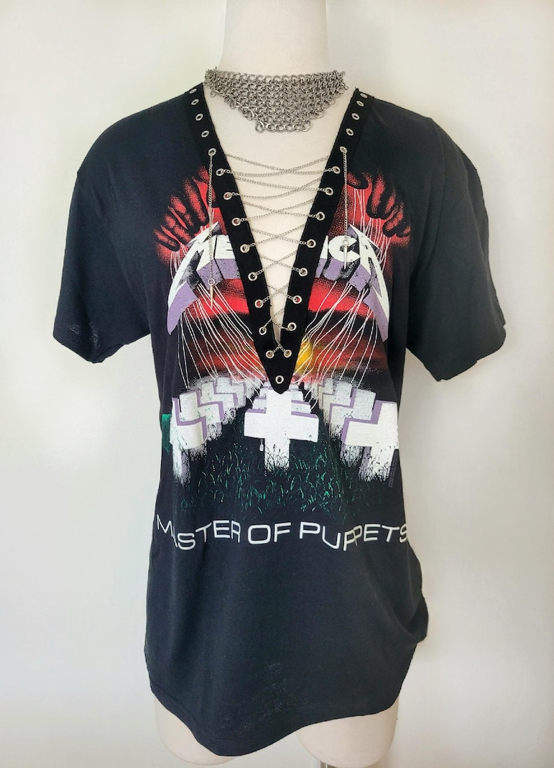 May include: Black t-shirt with a graphic of Metallica's "Master of Puppets" album cover. The shirt has a lace-up detail at the neckline with silver eyelets and a silver chain.