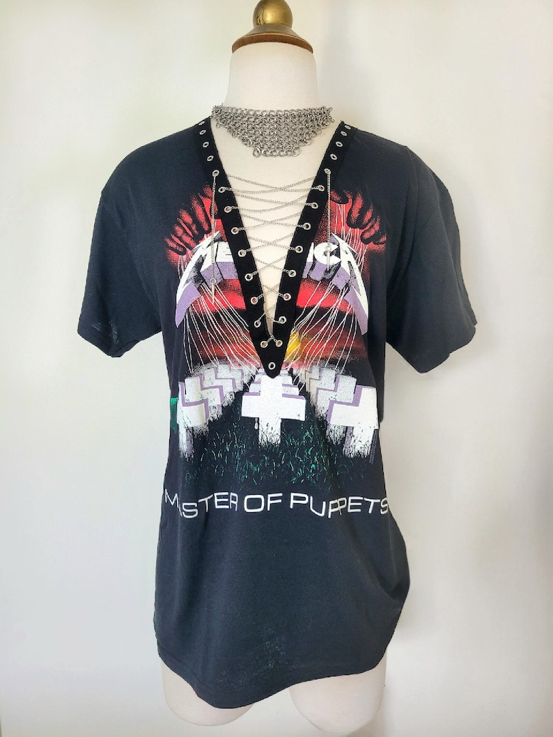 May include: Black t-shirt with a graphic of the band Metallica's "Master of Puppets" album cover. The shirt has a lace-up detail at the neckline with silver chain accents.