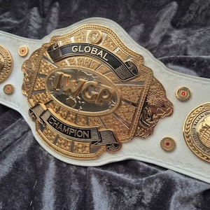Könnte beinhalten: Ein weißer Leder-Meisterschaftsgürtel mit einer großen, verzierten goldfarbenen Mittelplatte mit der Aufschrift GLOBAL IWGP CHAMPION. Der Gürtel enthält zwei runde goldfarbene Seitenplatten mit Weltkugel-Motiv und kleine rote Metallnieten.