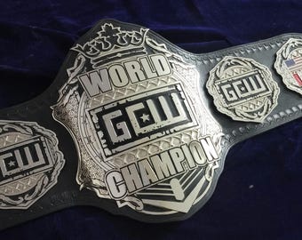 May include: A black wrestling championship belt with silver detailing. The belt displays the text "WORLD CHAMPION" and "GCW" on the main plate, and smaller plates with "GCW" and flags. The belt is made of leather.