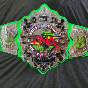 May include: A green and silver wrestling championship belt. The belt features the text "INTERNATIONAL CHAMPION" and "TNA" in red and black. The belt has a green strap and silver plates with globe and flag designs.