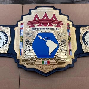 Puede incluir: Un cinturón de campeonato de lucha libre con una gran placa central chapada en oro y una incrustación hexagonal azul que muestra un mapa de América Latina. El texto CAMPEÓN LATINOAMERICANO está grabado sobre el mapa, flanqueado por dos placas laterales.