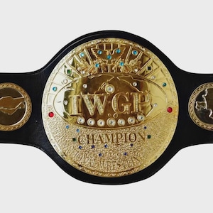 Könnte beinhalten: Ein Wrestling-Meisterschaftsgürtel mit einer großen, runden, goldfarbenen Mittelplatte mit der Aufschrift IWGP CHAMPION. Die Platte ist mit eingebetteten blauen, roten und klaren Kristallen, einem Adleremblem und einem schwarzen Lederriemen verziert.