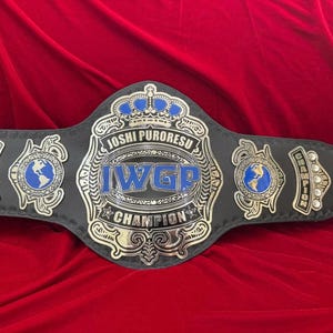 Könnte beinhalten: Ein schwarzer Wrestling-Meisterschaftsgürtel mit silbernen und blauen Akzenten. Der Gürtel trägt die Aufschrift "Joshi Puroresu" und "IWGP Champion". Er hat ein großes zentrales Emblem und kleinere seitliche Embleme.