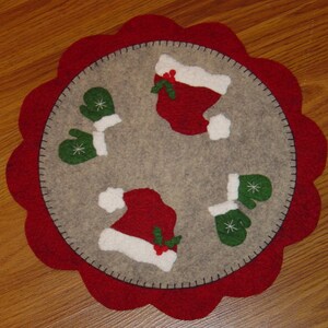 May include: A circular felt placemat with a red scalloped edge, featuring a Christmas theme. The grey centre has appliqued red Santa hats and green mittens with white trim. The hats and mittens are arranged in a circular pattern.