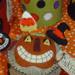 May include: A brown felt pincushion shaped like a smiling jack-o'-lantern with a black felt witch's hat on top. The pincushion has a candy corn pin on top and a white felt tag that says "Pinkeep".