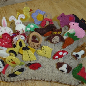 May include: A collection of colorful felt animal and nature-themed ornaments. The assortment includes a white bunny with pink ears, a yellow duck, a purple mouse, a red heart, a mushroom, and a brown owl. The ornaments are arranged on a gray felt base.