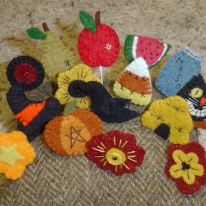 May include: A selection of handmade felt ornaments in diverse shapes, such as a red apple, watermelon slice, and black cat. The ornaments are crafted with stitched details and button accents, ideal for seasonal decorations and crafts.