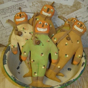 Puede incluir: Cuatro muñecas de calabaza hechas a mano con caras pintadas y detalles de campana. Las muñecas están vestidas con tela verde, marrón y naranja con lunares negros, blancos y naranjas. Las muñecas están sentadas en un tazón blanco con un borde verde.