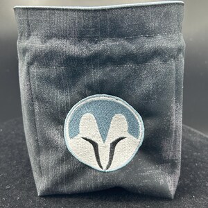 May include: A dark grey fabric pouch with a structured, boxy shape. The front features a circular embroidered patch with a light blue border, a white stylised helmet-like graphic, and black accents. The pouch has a simple, open-top design.