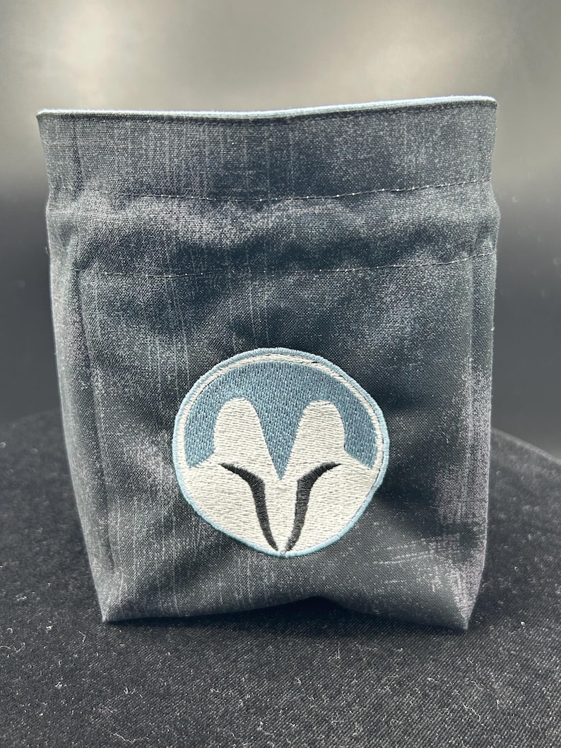 May include: A dark grey fabric pouch with a structured, boxy shape. The front features a circular embroidered patch with a light blue border, a white stylised helmet-like graphic, and black accents. The pouch has a simple, open-top design.