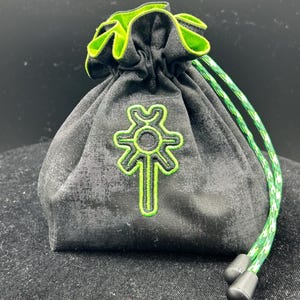 May include: Black drawstring bag with a green and white cord. The bag features a green embroidered design and a green trim around the opening. The bag is made of a textured fabric.