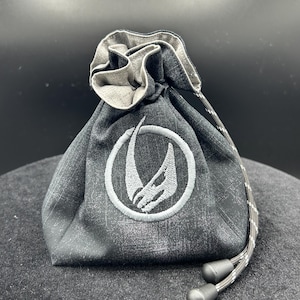 May include: A charcoal gray fabric drawstring pouch featuring a distressed texture and a circular embroidered emblem in light gray. The bag is cinched at the top with a matching patterned cord that includes two black plastic toggles at the ends.