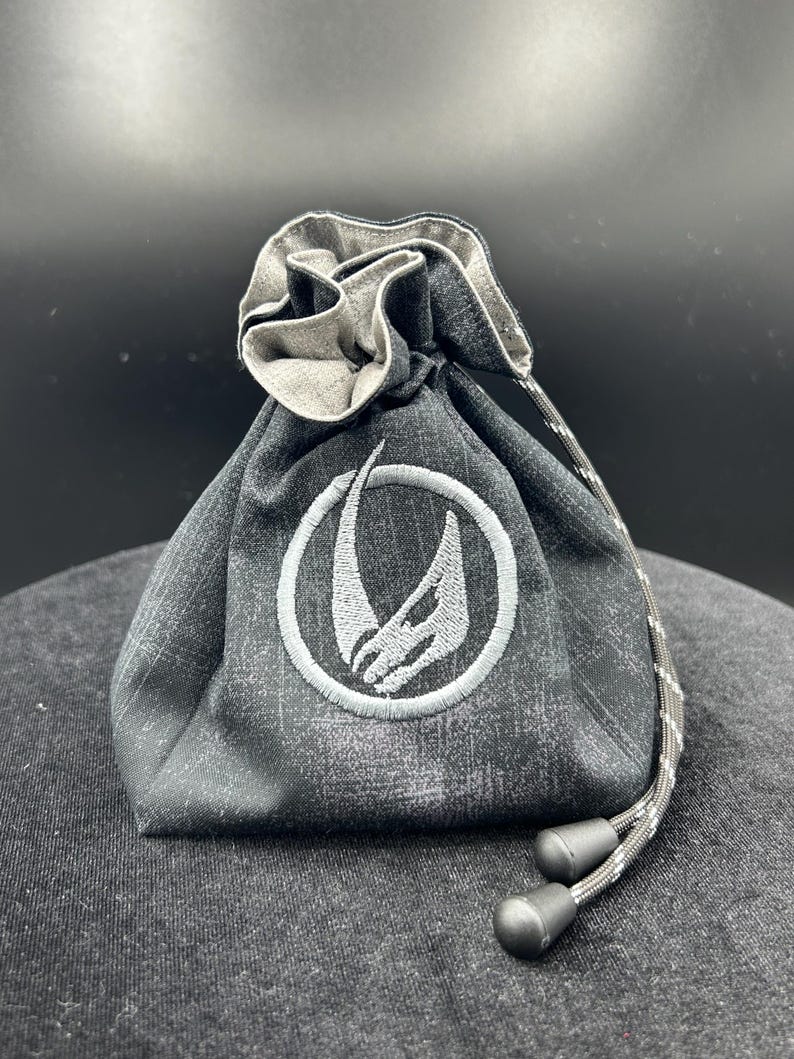 May include: A charcoal gray fabric drawstring pouch featuring a distressed texture and a circular embroidered emblem in light gray. The bag is cinched at the top with a matching patterned cord that includes two black plastic toggles at the ends.