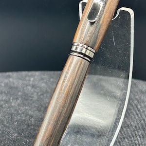 May include: A wooden pen with a dark brown finish and silver-colored metal accents. The pen is displayed on a clear acrylic stand against a dark background. The pen's design features a sleek, professional aesthetic.