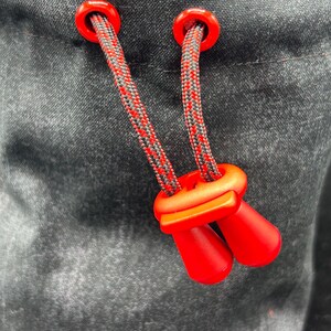 May include: A black fabric pouch featuring a drawstring closure with two red plastic eyelets and a matching red cord lock. The drawstring is made of a braided black and red cord that passes through the eyelets and secures with the red toggle.