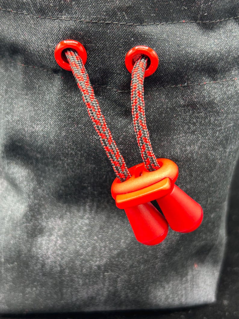 May include: A black fabric pouch featuring a drawstring closure with two red plastic eyelets and a matching red cord lock. The drawstring is made of a braided black and red cord that passes through the eyelets and secures with the red toggle.