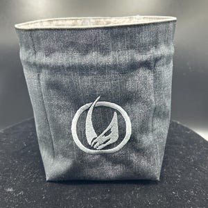 May include: A dark gray fabric storage bin with a textured, mottled appearance. The front features a circular embroidered emblem in light gray, depicting a stylized, curved horn or tusk shape enclosed within a thin, circular border.