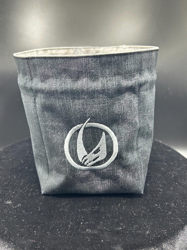 May include: A dark gray fabric storage bin with a textured, mottled appearance. The front features a circular embroidered emblem in light gray, depicting a stylized, curved horn or tusk shape enclosed within a thin, circular border.