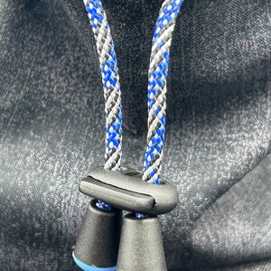 May include: A dark grey fabric surface featuring two metal eyelets through which a braided cord passes. The cord is patterned with blue, black, and white threads and is secured by a black plastic cord lock with two conical ends.