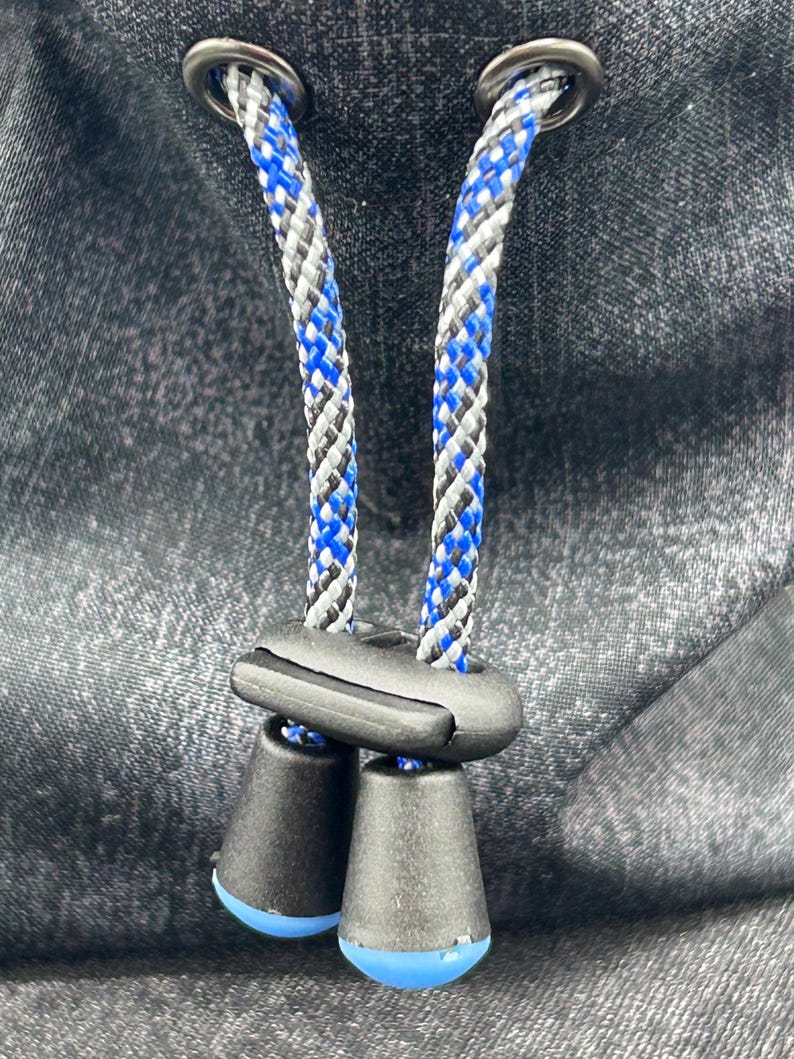 May include: A dark grey fabric surface featuring two metal eyelets through which a braided cord passes. The cord is patterned with blue, black, and white threads and is secured by a black plastic cord lock with two conical ends.
