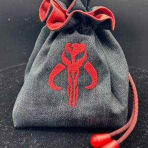May include: A charcoal gray fabric drawstring pouch featuring a red embroidered Mythosaur skull emblem on the front. The bag has a vibrant red interior lining and a matching red and gray cord with two solid red plastic toggles at the ends.