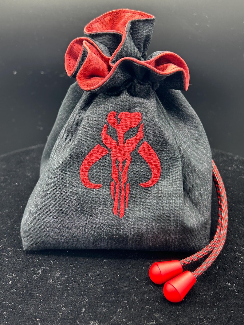 May include: A charcoal gray fabric drawstring pouch featuring a red embroidered Mythosaur skull emblem on the front. The bag has a vibrant red interior lining and a matching red and gray cord with two solid red plastic toggles at the ends.