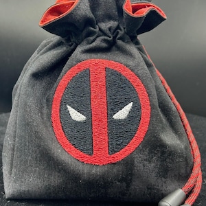 May include: Black drawstring bag with a red and black Deadpool logo embroidered on the front. The bag has a red interior and drawstring, and black plastic cord locks. The bag is made of a textured fabric.