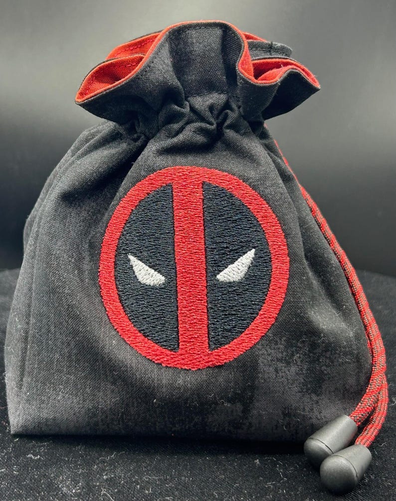 May include: Black drawstring bag with a red and black Deadpool logo embroidered on the front. The bag has a red interior and drawstring, and black plastic cord locks. The bag is made of a textured fabric.