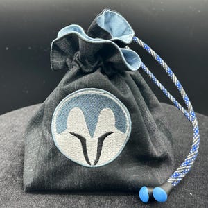 May include: A dark grey fabric drawstring pouch featuring a circular embroidered patch with a light blue and white stylised helmet design. The pouch has a light blue interior lining and a blue, grey, and black braided cord with two blue plastic toggles.