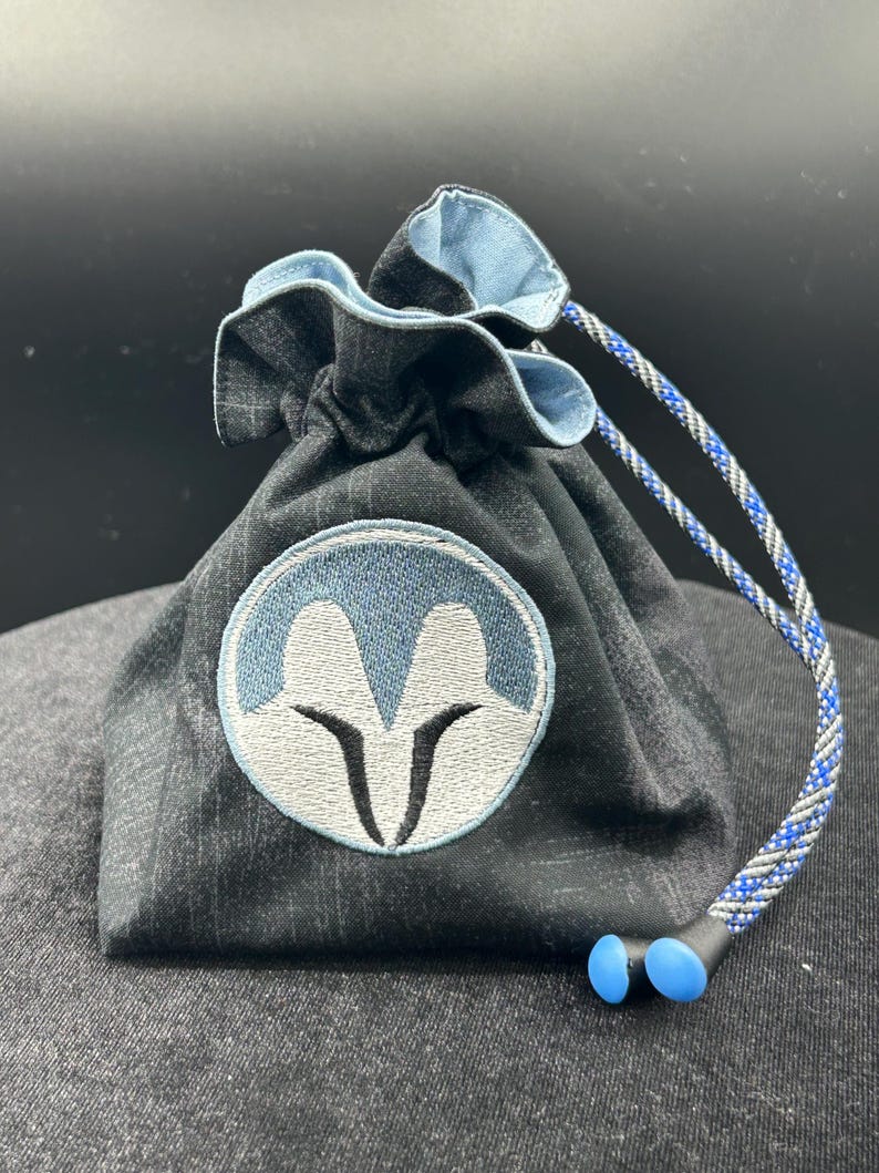 May include: A dark grey fabric drawstring pouch featuring a circular embroidered patch with a light blue and white stylised helmet design. The pouch has a light blue interior lining and a blue, grey, and black braided cord with two blue plastic toggles.