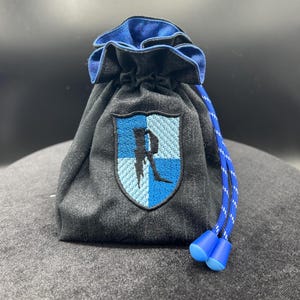 May include: A black drawstring bag with a blue interior and a blue and white emblem. The emblem features a stylized letter "R" and a crest design. The drawstring is blue and white, with blue plastic tips.