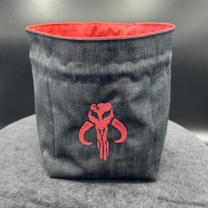 May include: A cylindrical fabric storage bin made of dark gray denim-like material with a red interior lining. The front features a prominent red embroidered Mythosaur skull emblem centered on the exterior surface of the container.