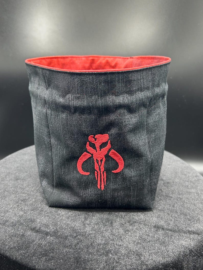 May include: A cylindrical fabric storage bin made of dark gray denim-like material with a red interior lining. The front features a prominent red embroidered Mythosaur skull emblem centered on the exterior surface of the container.