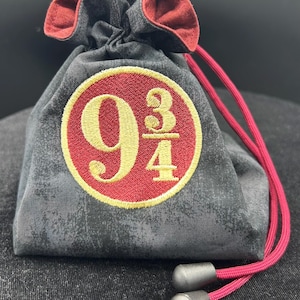 May include: A charcoal gray drawstring pouch with a red interior lining and red cord. The front features a circular embroidered patch with a red background and gold text reading 9 3/4. The drawstring ends with two dark gray cylindrical toggles.