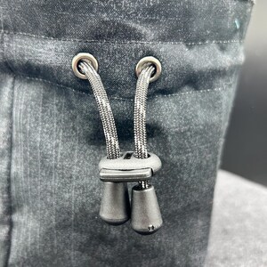 May include: A black fabric pouch featuring a drawstring closure with two silver-toned metal eyelets. The black cord passes through a black plastic cord lock with two dangling toggles, allowing for adjustable tightening of the bag opening.