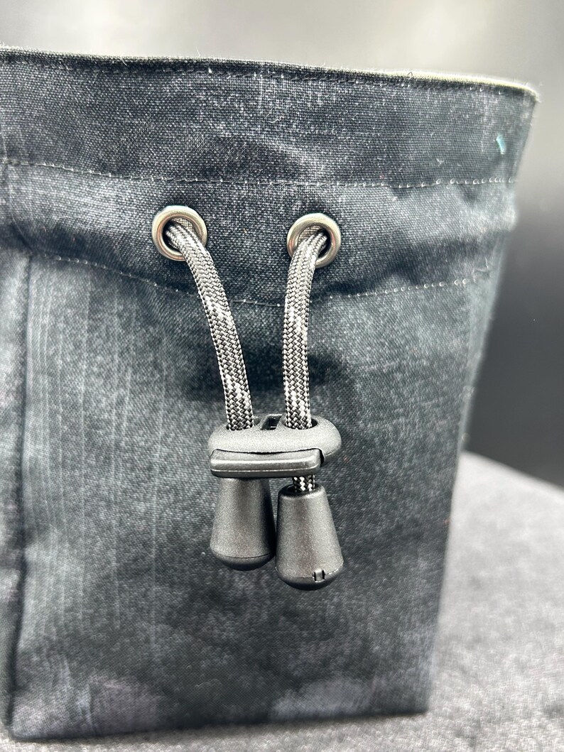 May include: A black fabric pouch featuring a drawstring closure with two silver-toned metal eyelets. The black cord passes through a black plastic cord lock with two dangling toggles, allowing for adjustable tightening of the bag opening.