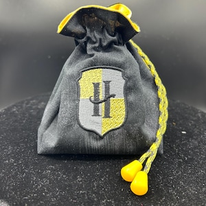 May include: Black drawstring bag with a yellow and gray cord and yellow plastic tips. The bag features a shield emblem with a yellow and gray design and the letter "H". The bag is made of a textured fabric.