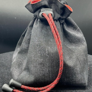 May include: A drawstring bag with a textured gray fabric exterior and a red interior. The bag features a red and black rope drawstring with black plastic tips and a black plastic cord lock. The bag is designed to be closed by pulling the drawstring.