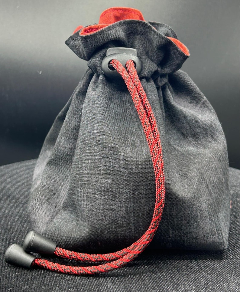 May include: A drawstring bag with a textured gray fabric exterior and a red interior. The bag features a red and black rope drawstring with black plastic tips and a black plastic cord lock. The bag is designed to be closed by pulling the drawstring.
