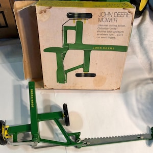 Puede incluir: Una segadora de juguete John Deere verde con una cuchilla plateada y ruedas negras. La caja muestra un diagrama de la segadora con el texto "John Deere Mower" y "Like-real cutting action. Cutterbar "knife" shuttles back and forth as wheels turn...won't cut small fingers."